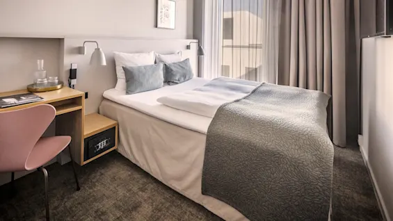 Siisti pedattu sänky harmailla ja valkoisilla vuodevaatteilla sijaitsee hotellihuoneessa, jossa on pehmeä valaistus. Sängyn vieressä on työpöytä tuoleineen, ja lähellä on ikkuna.