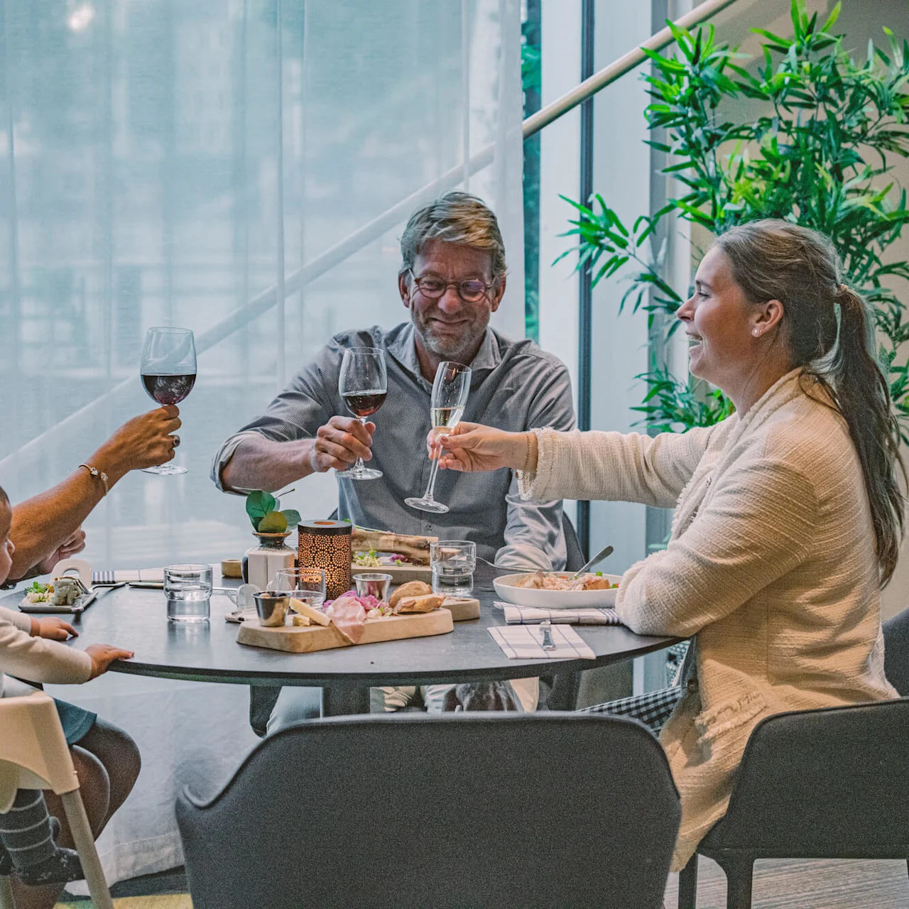A group of three adults and a child sit around a dining table, toasting with wine glasses, in a cozy restaurant with lush greenery and soft lighting.