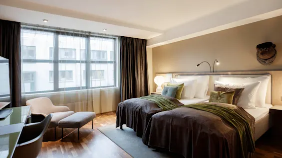 A neatly arranged hotel room features two beds with brown and green bedding, a seating area with armchair and ottoman, near a large window with sheer curtains.