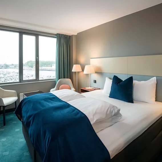 Et moderne hotellrom med en pent oppredd seng og blå detaljer er opplyst av to sengelamper. Et stort vindu viser en marina med båter og grøntområde.