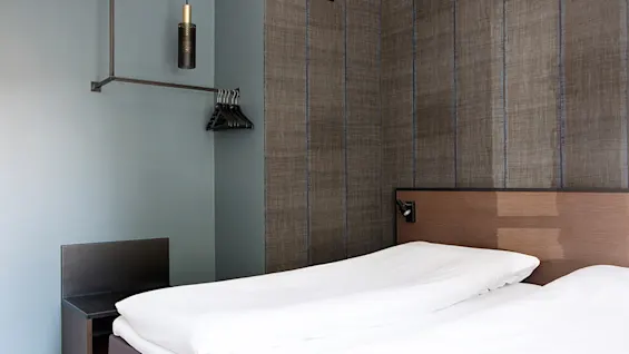 En pænt redt dobbeltseng med hvidt sengetøj står op ad et træhovedgærde; en tom metalgarderobe med bøjler er monteret på en væg i et moderne, minimalistisk rum.