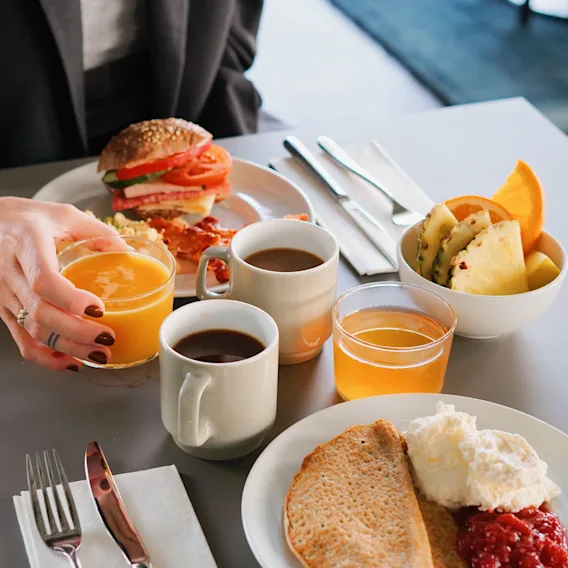 En hånd strekker seg etter et glass appelsinjuice på et bord dekket med en deilig frokostbuffé inkludert en sandwich, bacon, kaffe, frukt og pannekaker.