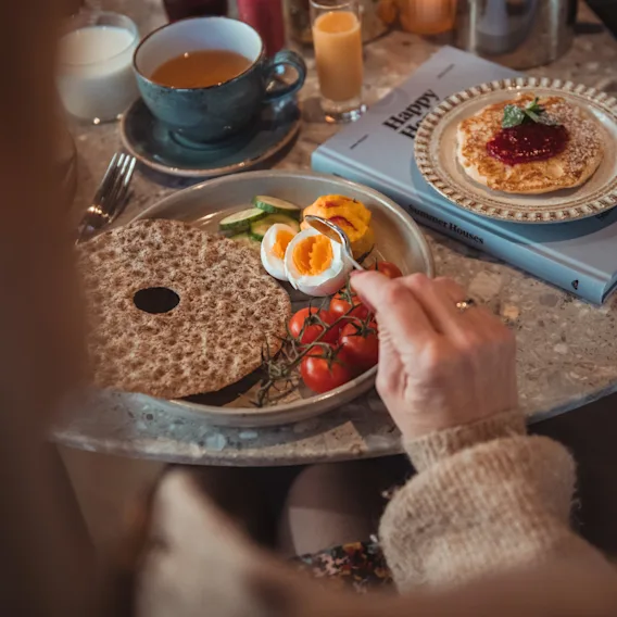 En person nyter frokost med knekkebrød, egg, tomater og en pannekake, med en bok 'Happy H' i nærheten.