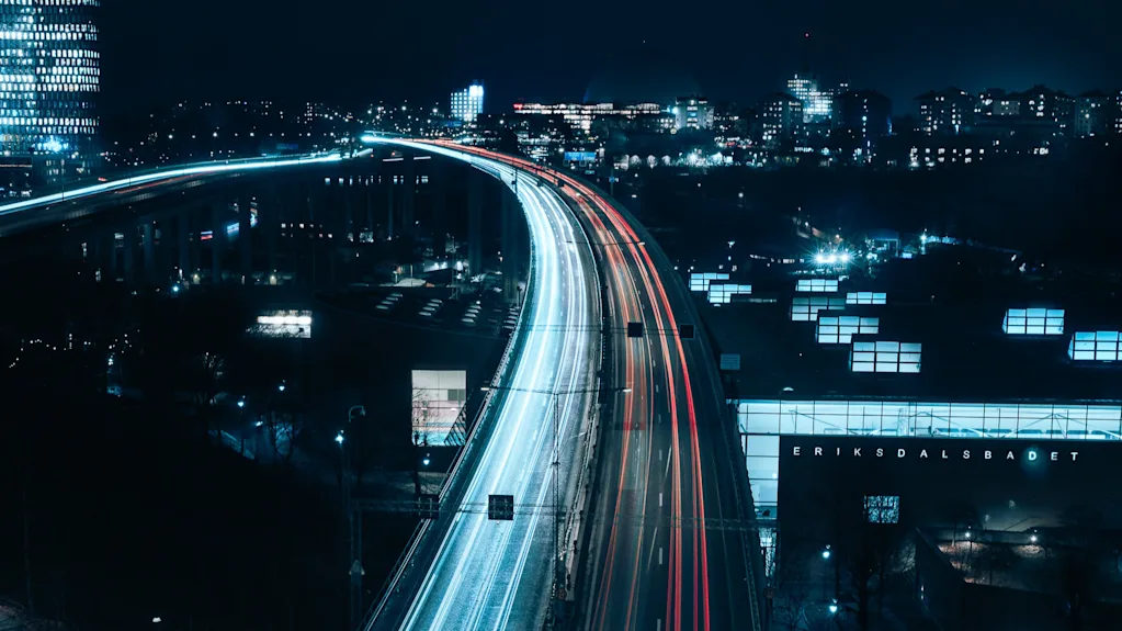 Gatubelysning skapar strimmor över en motorväg på natten, med stadens byggnader upplysta i bakgrunden. Text: "ERIKSDALSBADET".