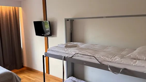 Bunk beds with neatly folded white sheets and towels, placed in a room with wooden floors. A wall-mounted TV is visible near a window covered by dark curtains.