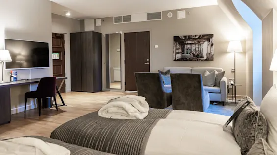 A hotel room features two beds with folded robes, a flat-screen TV on a desk, a mirror, and a seating area with a sofa and chairs, enhancing a cozy atmosphere.