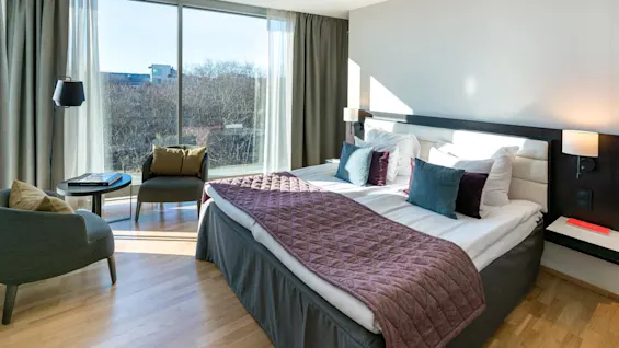 En pent sengered dobbeltseng med fargerike puter og et lilla teppe står i et lyst hotellrom. Stoler og et bord er plassert ved store vinduer med utsikt over trær.
