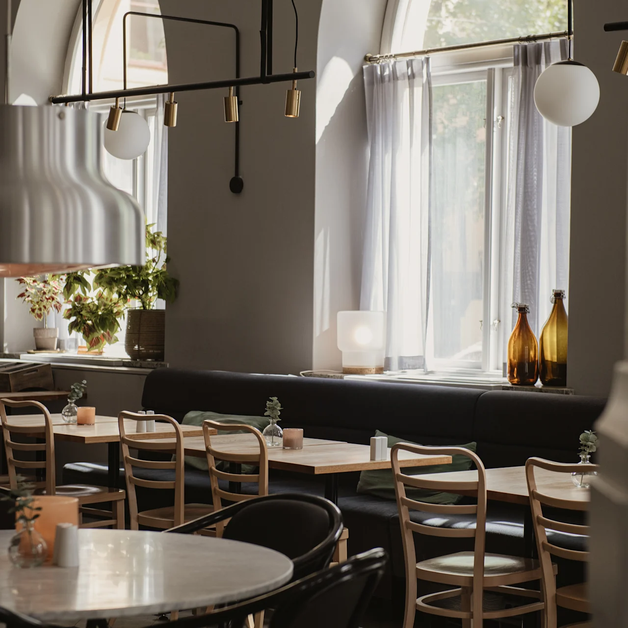 Ett långt matbord med trästolar och små växter står i ett mjukt upplyst, modernt café. Stora fönster med gardiner tillåter naturligt ljus och framhäver dekorativa flaskor och växter.