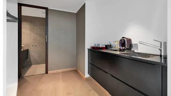 Coffee machine on dark countertop; surrounded by cups, glasses, and a sink; located in a modern room with wood floors and beige walls, next to an open bathroom door.