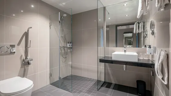 Moderne badeværelse med væghængt toilet, indglasset bruser og vask på sort bordplade. Beige fliser dækker væggene; håndklæder og toiletartikler er pænt arrangeret.