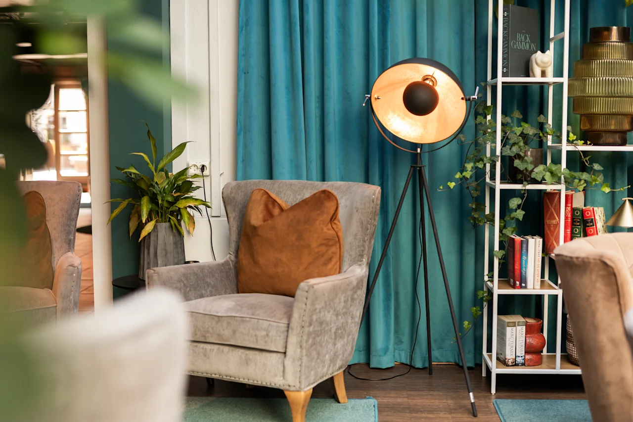 Fåtölj med brun kudde står i rum med blå gardiner. Lampa på stativ står bredvid. Bokhylla med böcker och dekorativa föremål fyller hörnet. Lummig växt kompletterar den mysiga platsen.