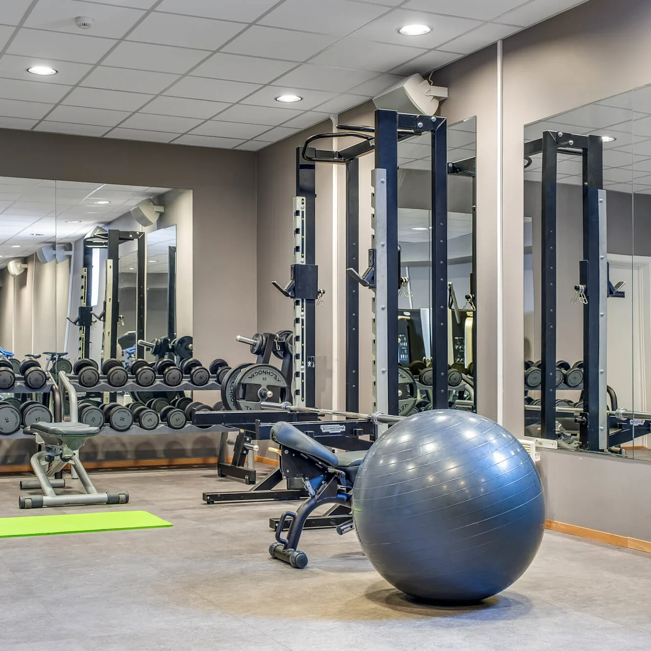 Exercise equipment in a gym, including weights, mats, and a stability ball, with mirrors on the wall.