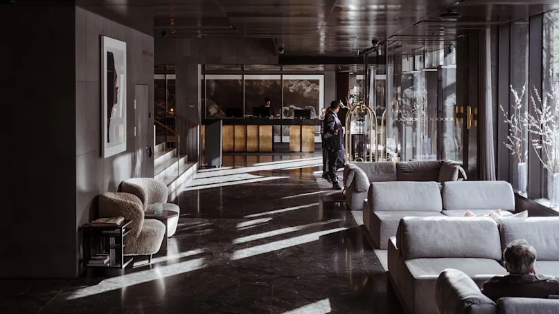 A hotel lobby with a reception desk; a person checks in. Soft couches and armchairs fill the sunlit space, with modern decor and window views.