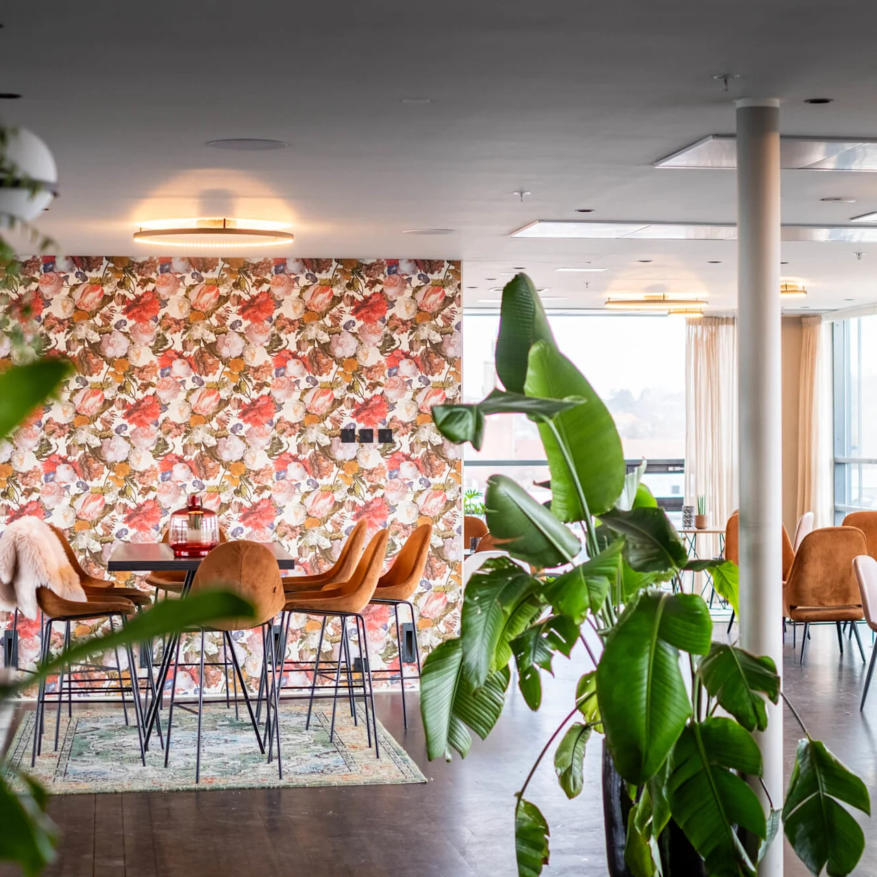 A dining area features brown chairs around tables against a floral-patterned wall. Large potted plants decorate the space, which is well-lit by natural light through nearby windows.