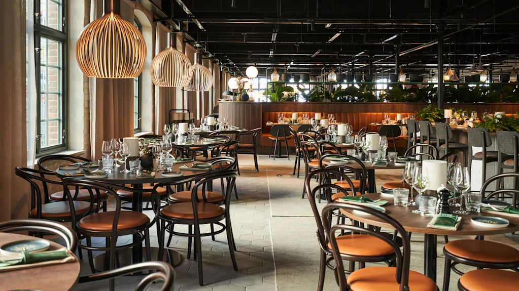 Bord dukade med glas och tallrikar, omgivna av trästolar i en rymlig restaurang med stora fönster och hängande lampor, vilket skapar en elegant atmosfär.