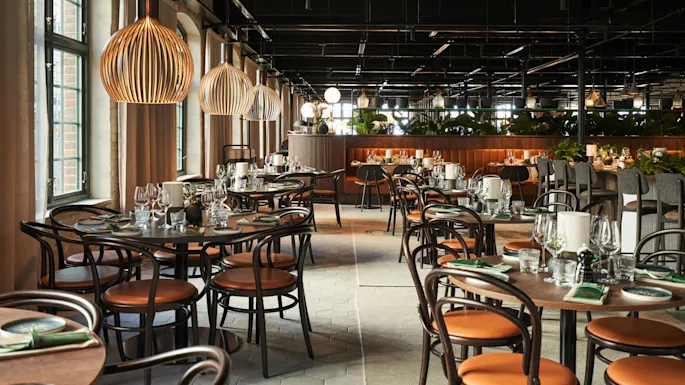 Bord dekket med glass og tallerkener, omgitt av trebarnstoler i en romslig restaurant med store vinduer og hengende lamper, som skaper en elegant atmosfære.