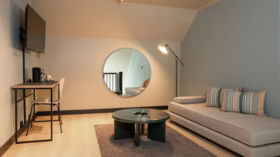 Ett bekvämt och stilfullt hotellrum med en dagbädd, ett litet skrivbord med TV och en framträdande cirkulär spegel.