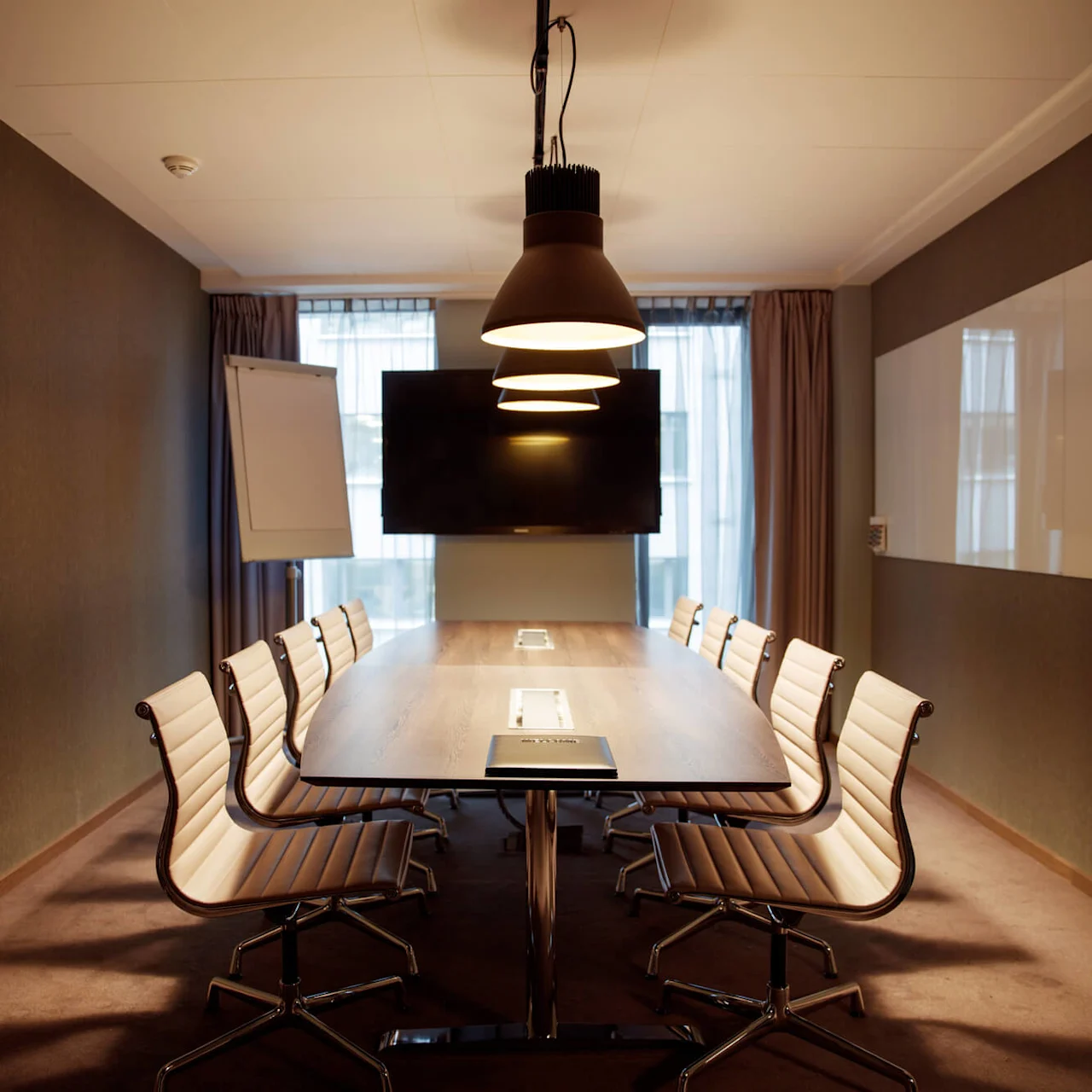 A conference room with a long wooden table, surrounded by modern chairs, is lit by pendant lamps. A large screen and a flip chart stand are at the far end.