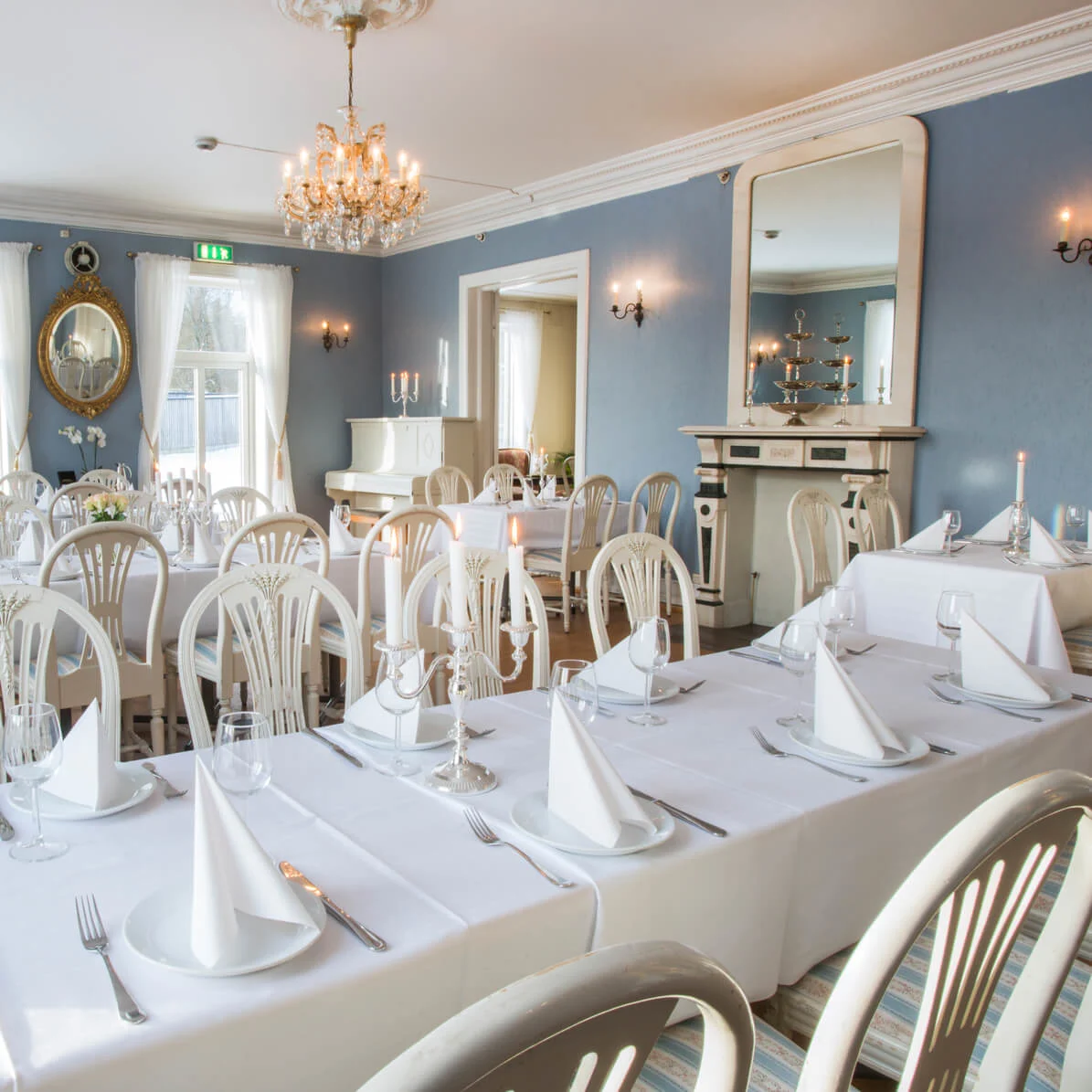 Elegant matsal med dukade bord, vita stolar och en kristallkrona. Blå väggar och klassisk inredning skapar en formell atmosfär.