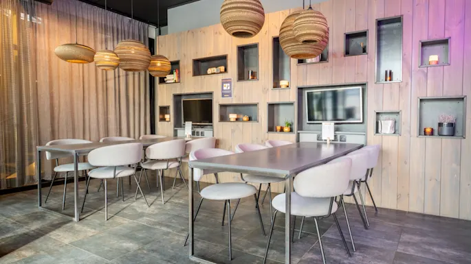 Chairs surround tables in a modern dining area with wooden panels, built-in shelves, two TVs, pendant lights, and a semi-transparent curtain. The setting appears stylish and contemporary.