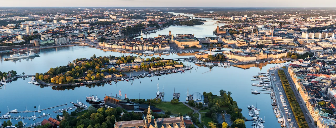 A historic building is surrounded by lush trees and water, nestled within an urban landscape. Numerous boats are docked along the waterfront, under a vast, clear sky.