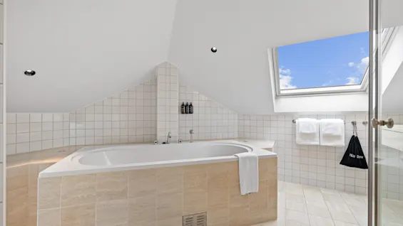 Lyst, flislagt bad på Home Hotel Gabelshus med stort badekar under takvindu som viser blå himmel. Hårfønerpose henger i nærheten.