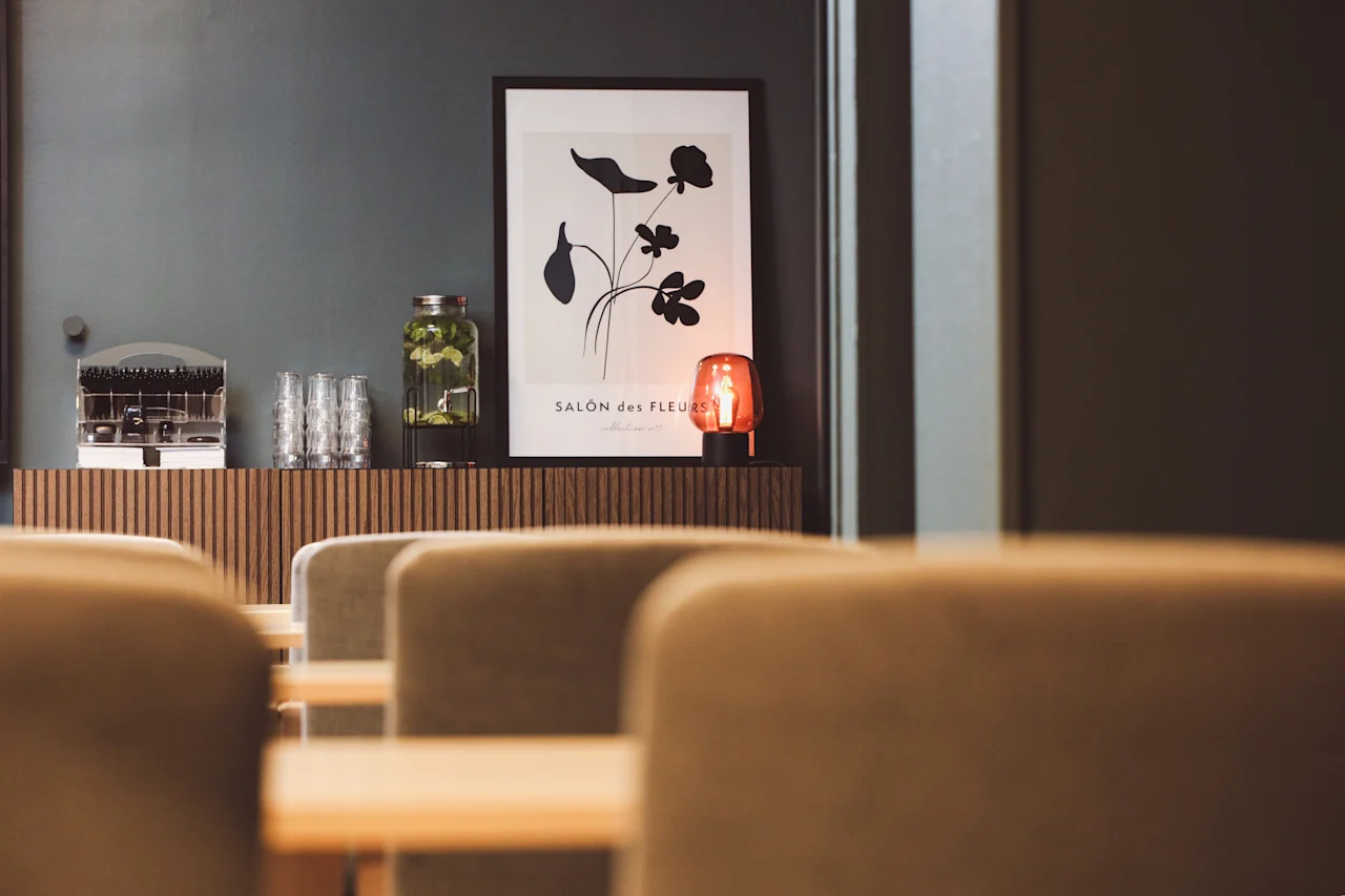 En inramad botanisk affisch hänger ovanför ett skåp med en burk grönt, glas och en röd lampa i en mysig matplats. Texten lyder: "SALON des FLEURS."