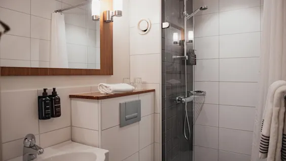 A clean and modern bathroom at Home Hotel Kung Oscar, featuring a sink, mirror, and a walk-in shower with fresh towels.