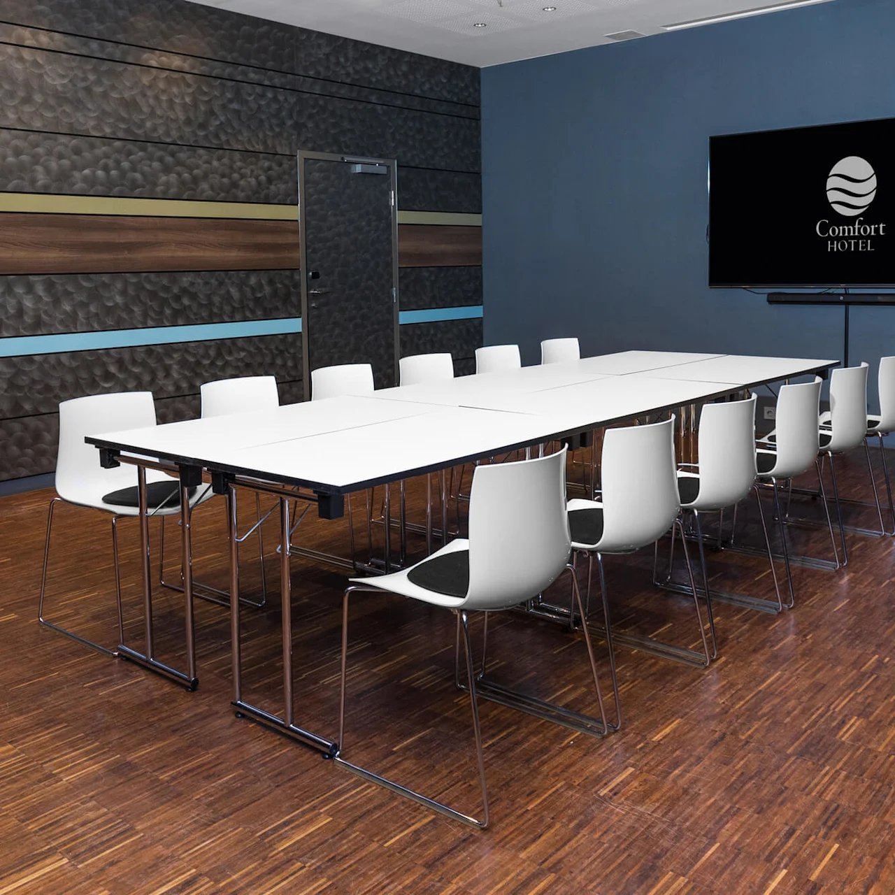 White conference table surrounded by white chairs in a meeting room with wooden floors. "Comfort Hotel" logo displayed on a large screen in the background.