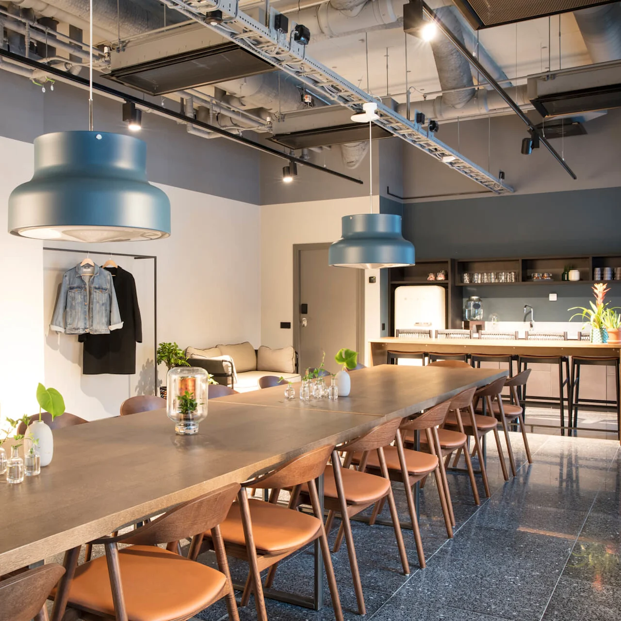 A long wooden table is surrounded by chairs in a modern, well-lit room. Blue pendant lights hang above, with a kitchenette and coat rack in the background.