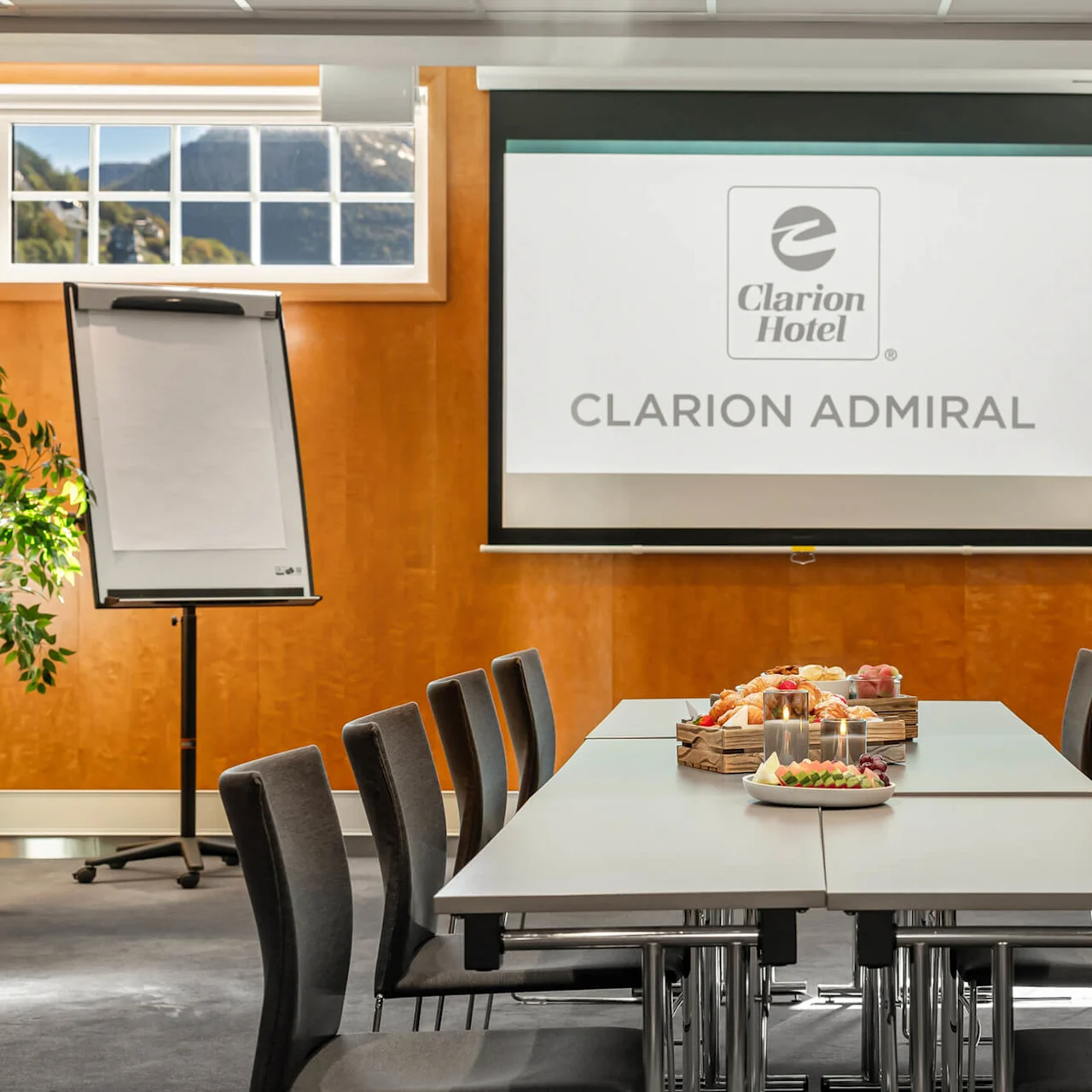 Konferencelokale med stole omkring et bord med snacks, mod en baggrund af en stor skærm med teksten "Clarion Hotel CLARION ADMIRAL"; flipchart og vinduer er i nærheden.