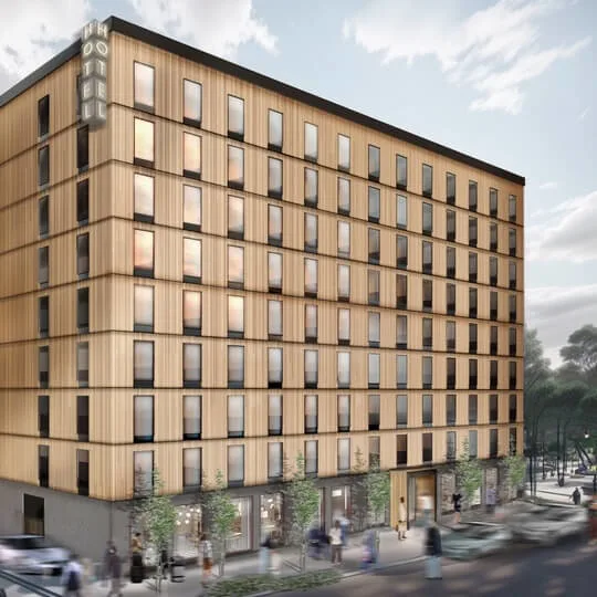Rakennus seisoo korkeana puujulkisivulla, merkintä "HOTEL." Ihmisiä ja autoja liikkuu puiden reunustamaa katua pitkin. Taivas on pilvinen pehmeän valon siivilöityessä läpi.