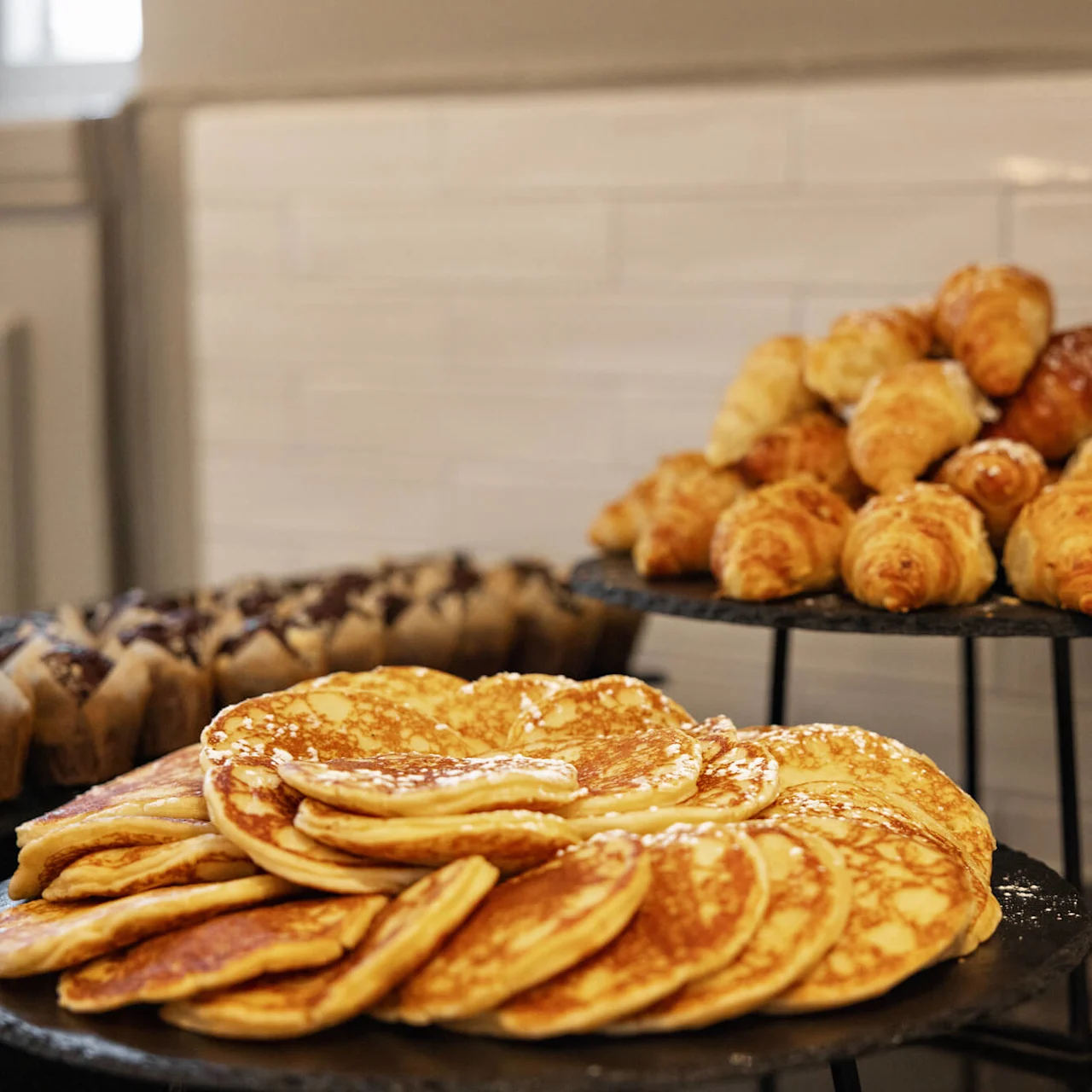 Pandekager og croissanter er pænt arrangeret på metalbakker, sammen med muffins i en hyggelig indendørs setting. Friske æbler tilføjer et livligt præg i en skål i nærheden.