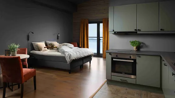 A cozy bedroom-kitchen space features a neatly made bed next to a small dining area with two chairs. The kitchenette has green cabinets, with an oven below. Large windows overlook a scenic view.