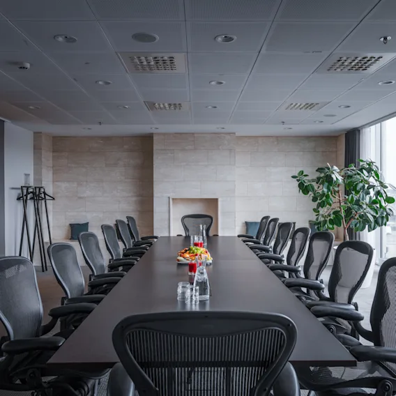 A large wooden conference table has several ergonomic chairs surrounding it in a modern meeting room with glass walls. A coat rack stands in the corner beside an entryway.