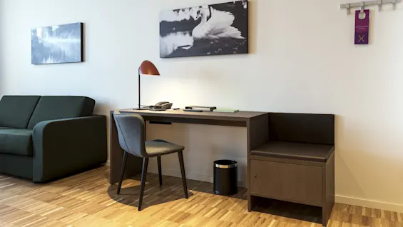 A wooden desk with a chair holds office supplies beneath a desk lamp. Nearby, a green sofa sits against the wall, decorated with nature-themed artwork. Text: "PLEASE DO NOT DISTURB".