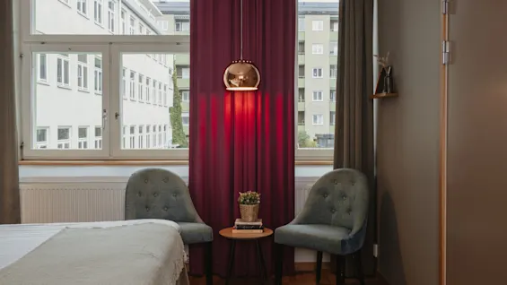 Home Hotel Uppsala room with two chairs, a table with books and a plant, and a bed. A pendant light casts a warm red glow.