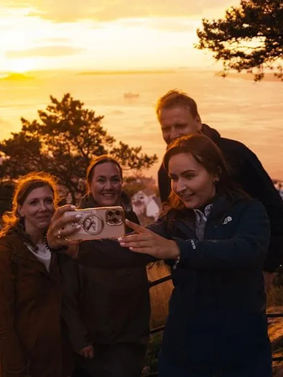 En gruppe venner tar en selfie med en vakker solnedgang over en kystby og vann i bakgrunnen.