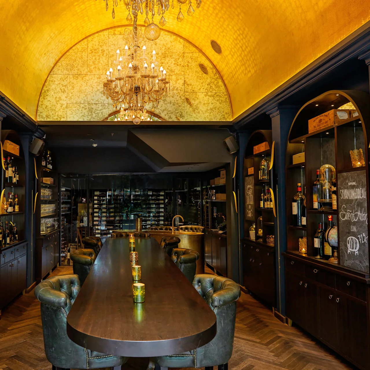 Elegant dining table surrounded by green leather chairs; shelves with bottles on both sides. Warm golden ceiling with chandelier, dark walls, and wooden floor. Blackboard with the text: "Sprøde Skagenskagernerau" and "Oste variation DKK 112."