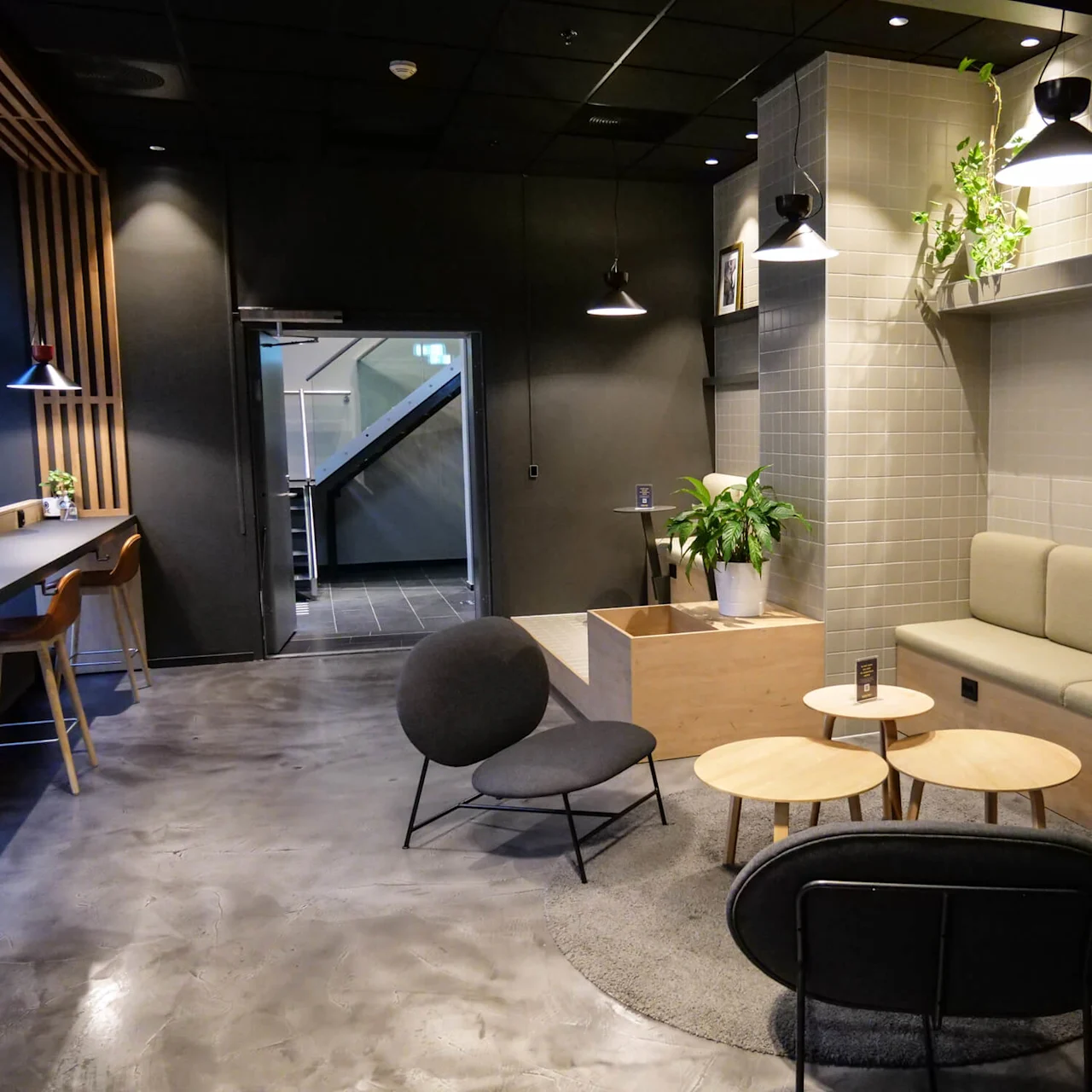 A modern lounge features cushioned seating and round tables, surrounded by green plants and soft lighting. It opens to a hallway with visible staircases.
