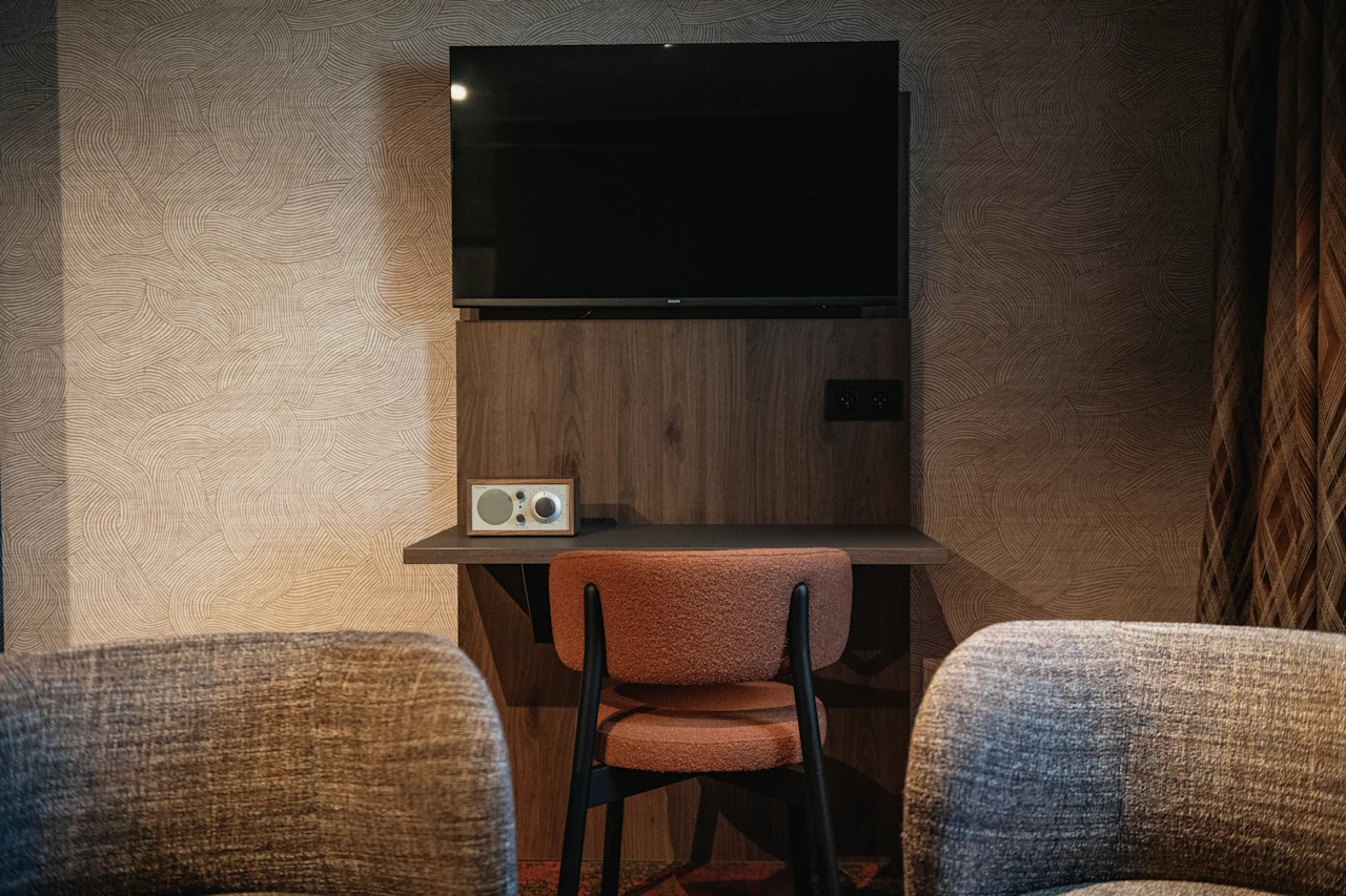 A hotel room corner with a wall-mounted TV above a desk, a chair, and two armchairs, ready for use.