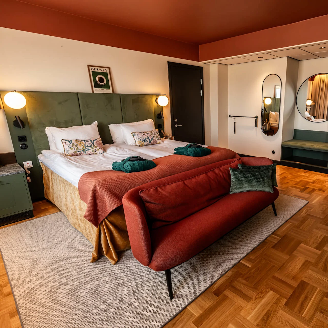 Twin beds with plush pillows and robes sit beside a green headboard. A red couch faces them, with wooden flooring and minimalist decor creating a cozy atmosphere.