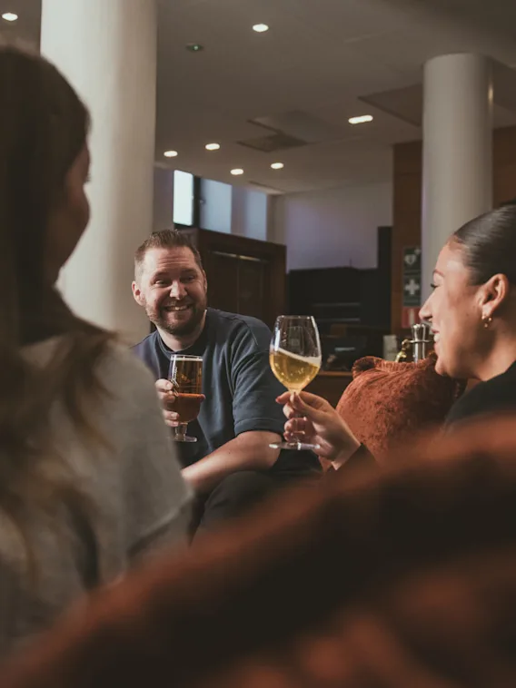 Tre personer sitter i en mysig lounge, njuter av drycker och ler under en konversation. En lampa ger varm belysning och växter ger en touch av grönska i bakgrunden.