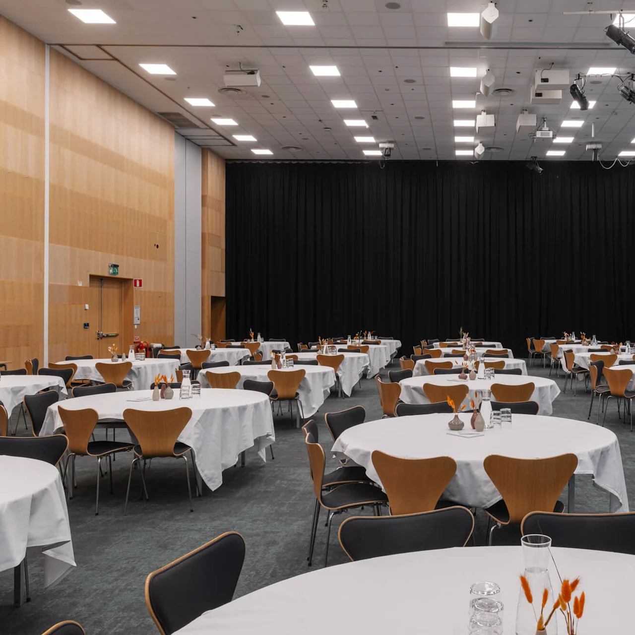 Runda bord täckta med vita dukar är arrangerade i ett stort konferensrum. Trästolar står runt borden. En scen med en skärm syns i bakgrunden.