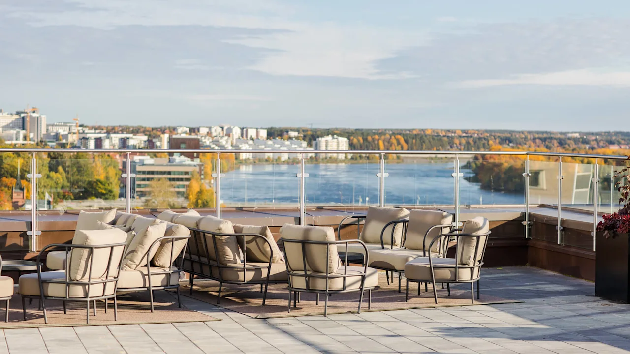 Komfortable utemøbler står ubrukt på en takterrasse, med utsikt over en naturskjønn elv og bybildet med trær i høstfarger i det fjerne under en klar himmel.
