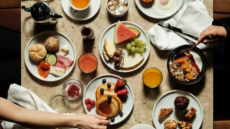 En livlig morgenmadsbuffet med pandekager, frisk frugt, æg, bacon og bagværk, hvor hænder rækker ud efter mad.