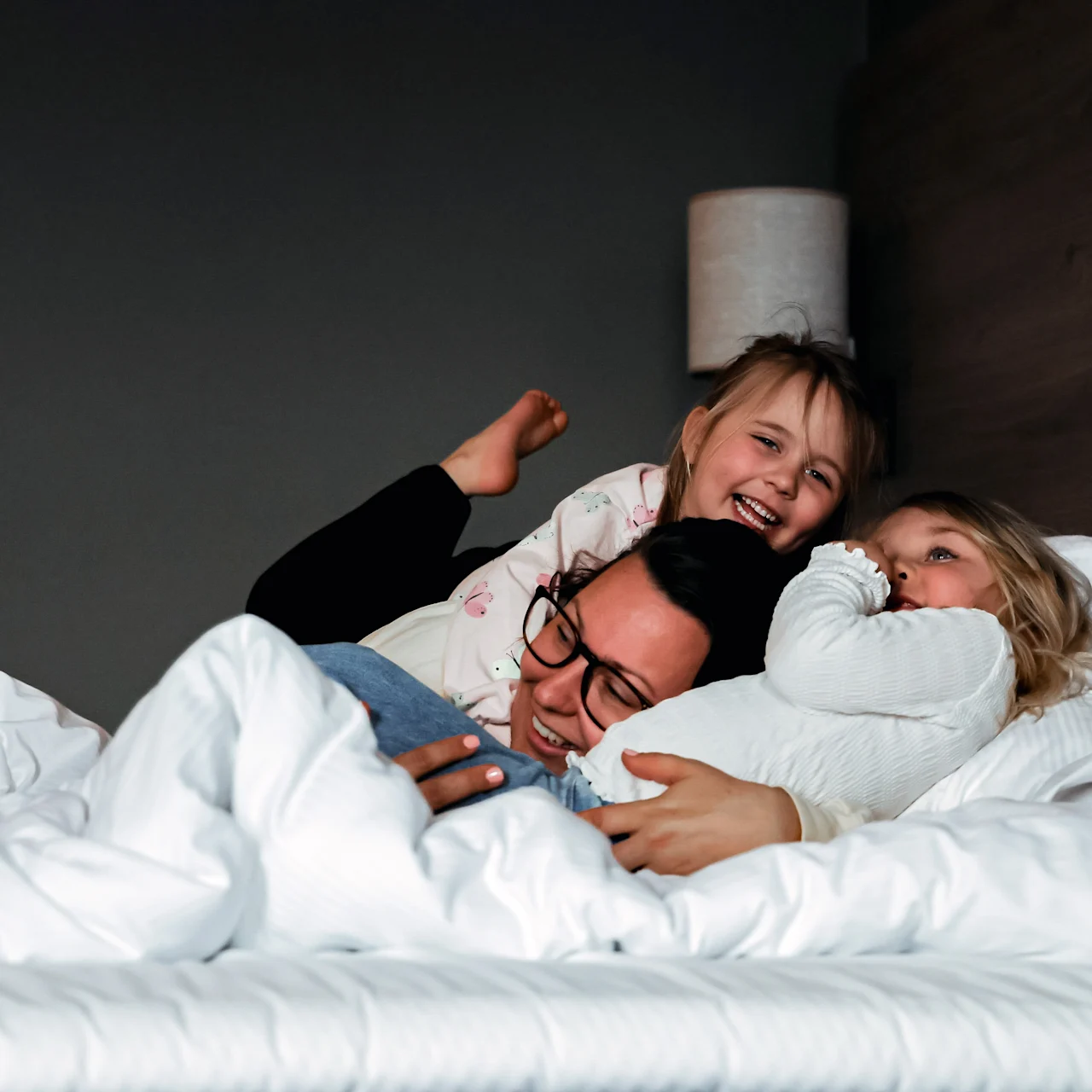 A person lies in bed with two children playfully embracing them, all smiling. The bed is covered with white sheets in a cozy bedroom setting.