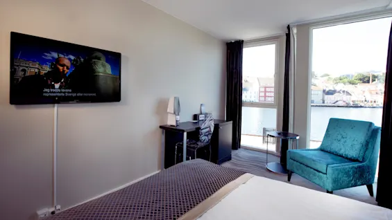 A modern hotel room with a mounted TV displaying a show, and a view overlooking a waterway and colorful buildings.