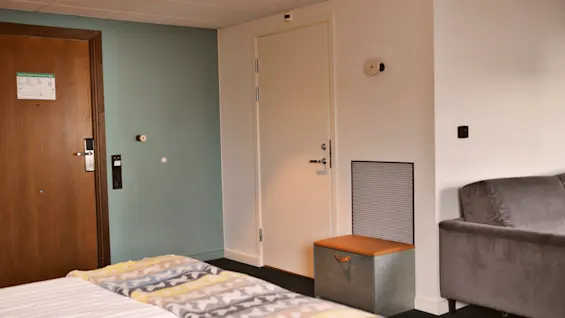 Et hotelværelse med en pænt redt seng, farverige mønstrede tæpper, en trådør med en seddel, en lukket lys dør og en grå sofa mod væggen.