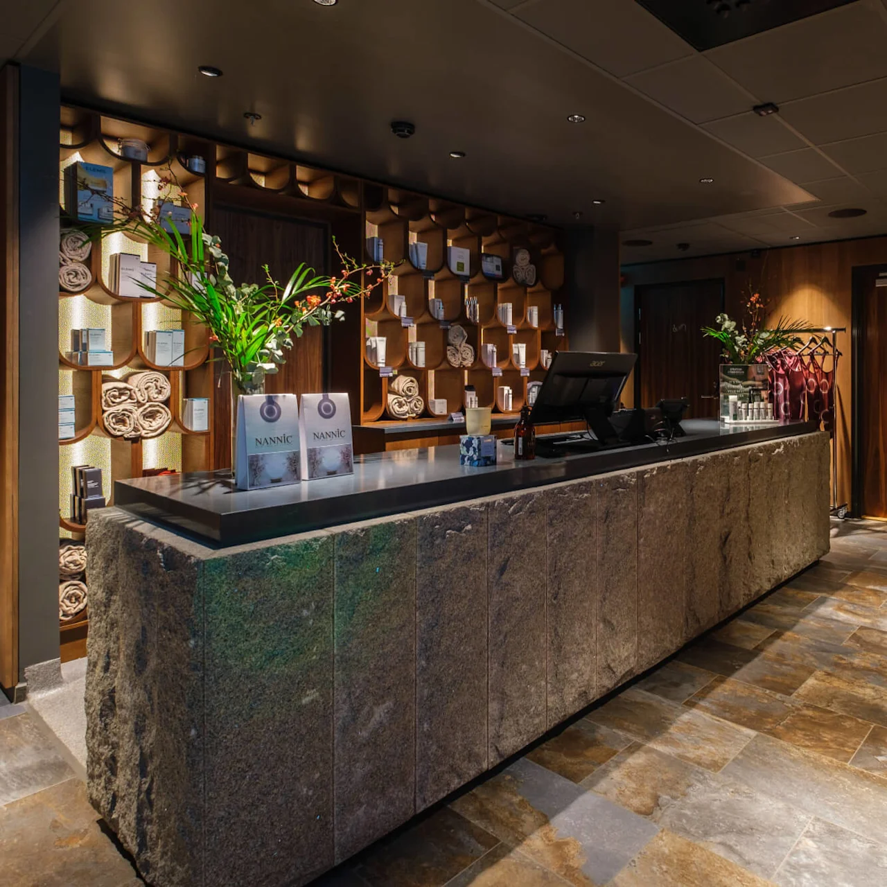En granitdisk visar broschyrer och en blomvas; bakom, hyllor med ihoprullade handdukar och produkter. Miljön liknar ett spa med dämpad belysning. Texten lyder "DRESSING ROOM" på en dörr.